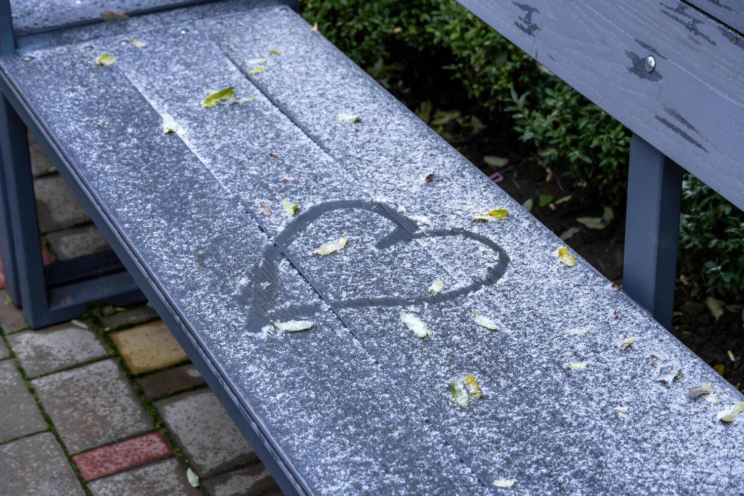 A heart shape is etched in the snow covering a bench, with fallen leaves scattered around it, creating a serene winter atmosphere photo
