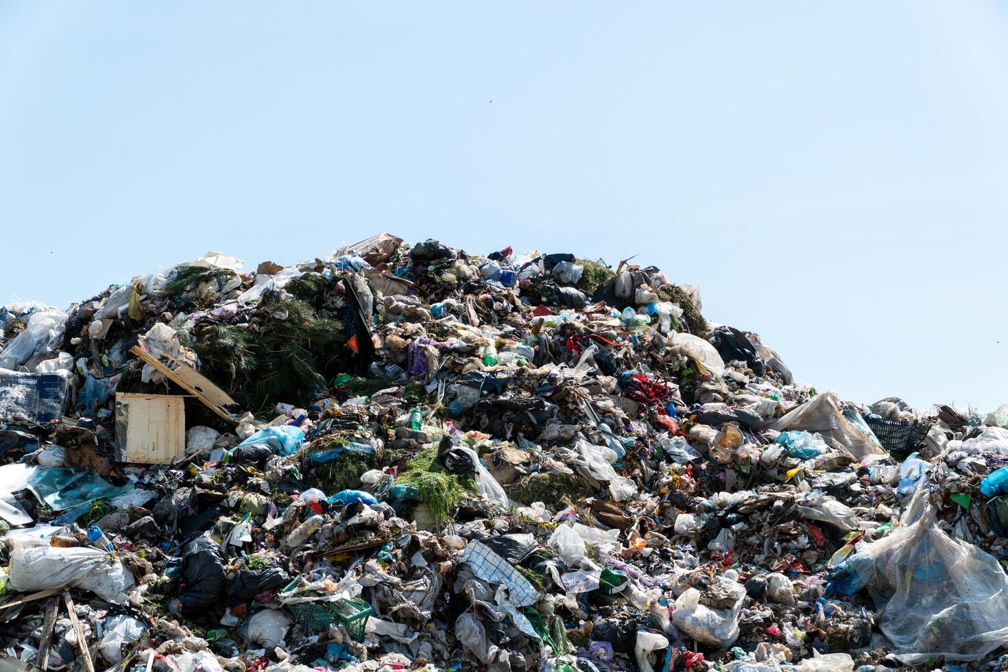 Piles of plastic and various waste materials fill the landfill under a clear blue sky. Littered debris and garbage are visible throughout photo