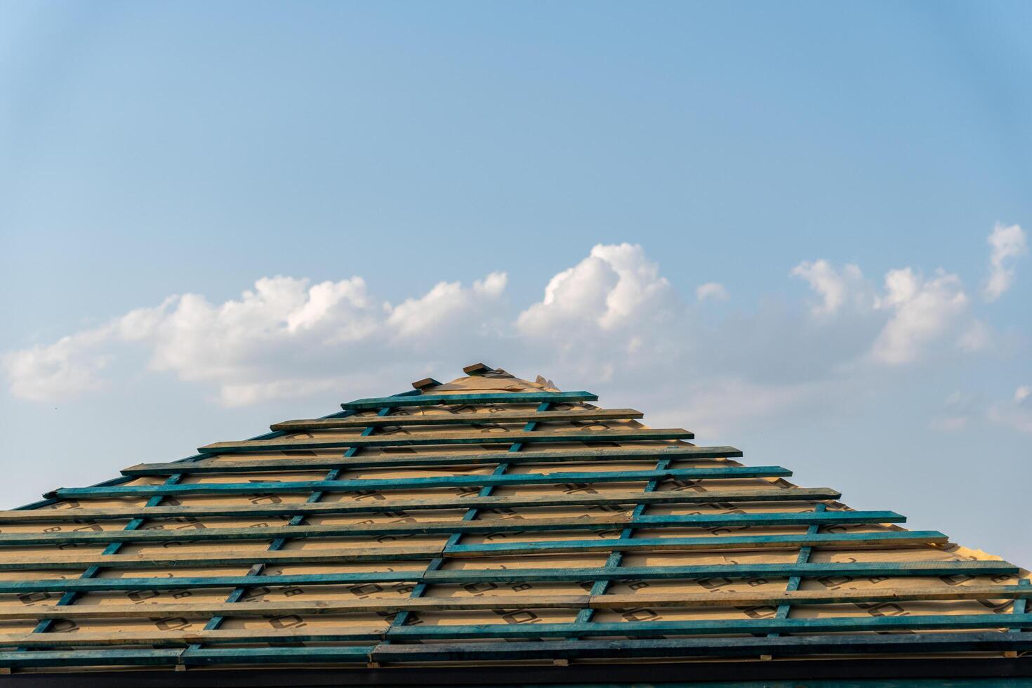 Workers are installing wooden beams on a roof, with a clear blue sky and fluffy clouds above, creating a serene construction setting photo