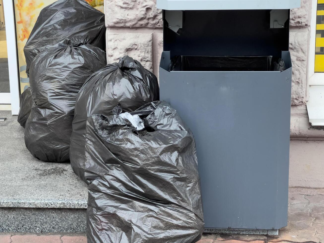 Black garbage bags are piled next to a public trash bin outside a storefront, creating an unkempt appearance in the urban environment photo