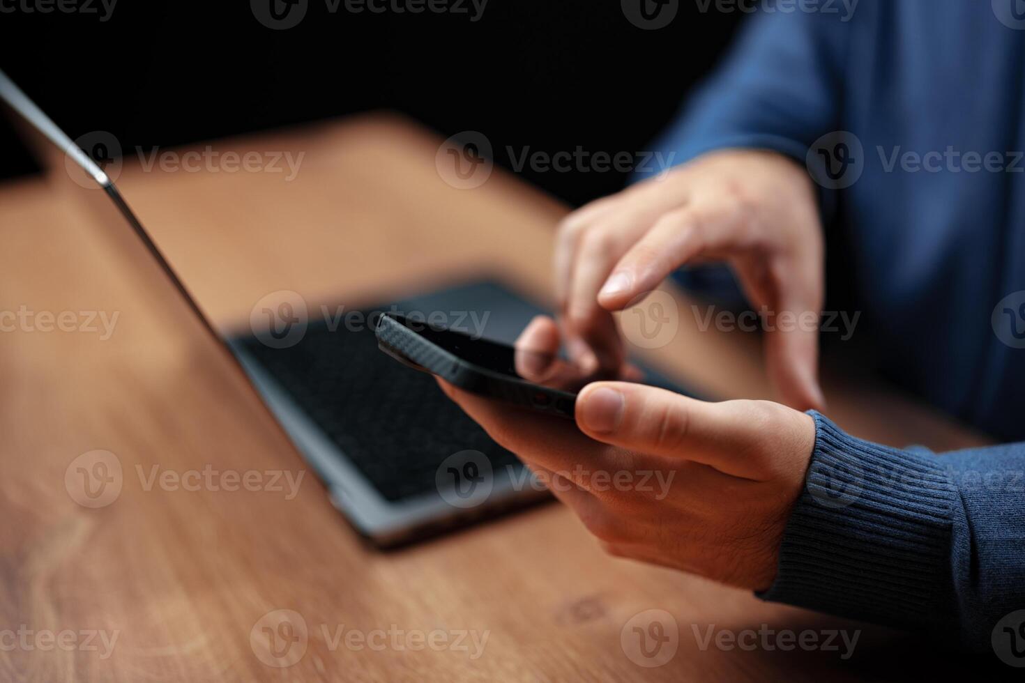 Person using smartphone while sitting at wooden desk with laptop during evening hours in a cozy setting photo