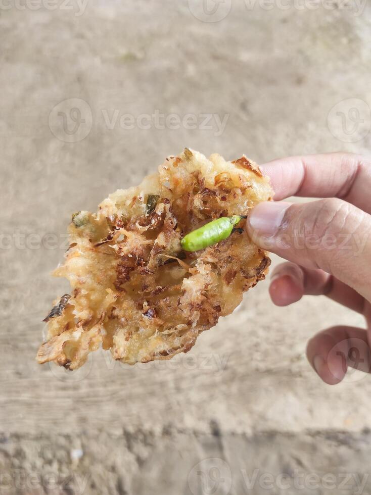 Close-up shot of the right hand holding fried fritters with green chilies. A crispy fried food made from a mixture of flour and carrots, bean sprouts, and cabbage. Indonesian food with negative space. photo