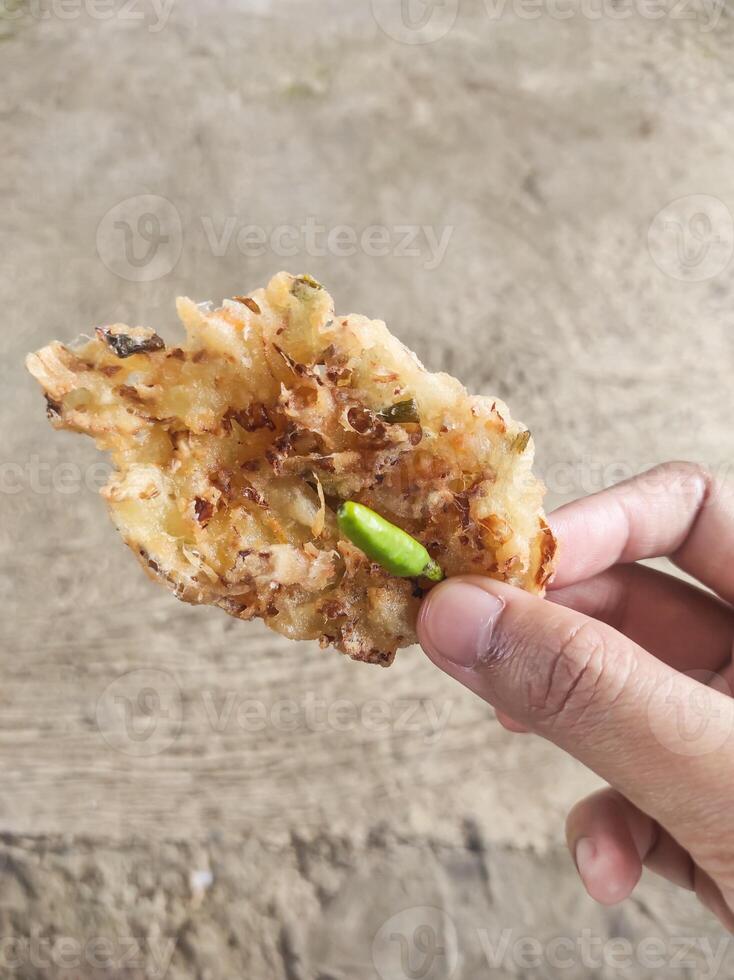 Close-up shot of the right hand holding fried fritters with green chilies. A crispy fried food made from a mixture of flour and carrots, bean sprouts, and cabbage. Indonesian food with copy space. photo