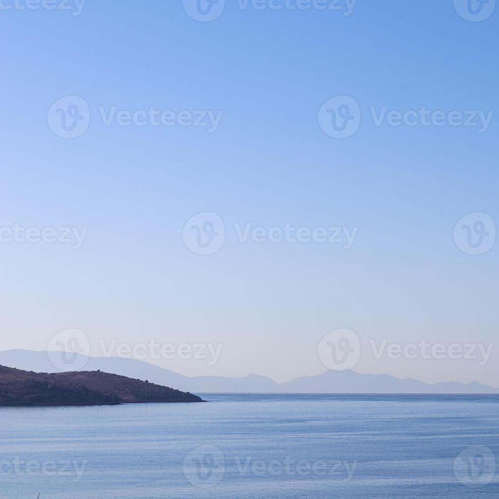 A tranquil blue seascape with a dark coastline in the foreground, and hazy distant mountains fading into a clear, gradient sky. photo