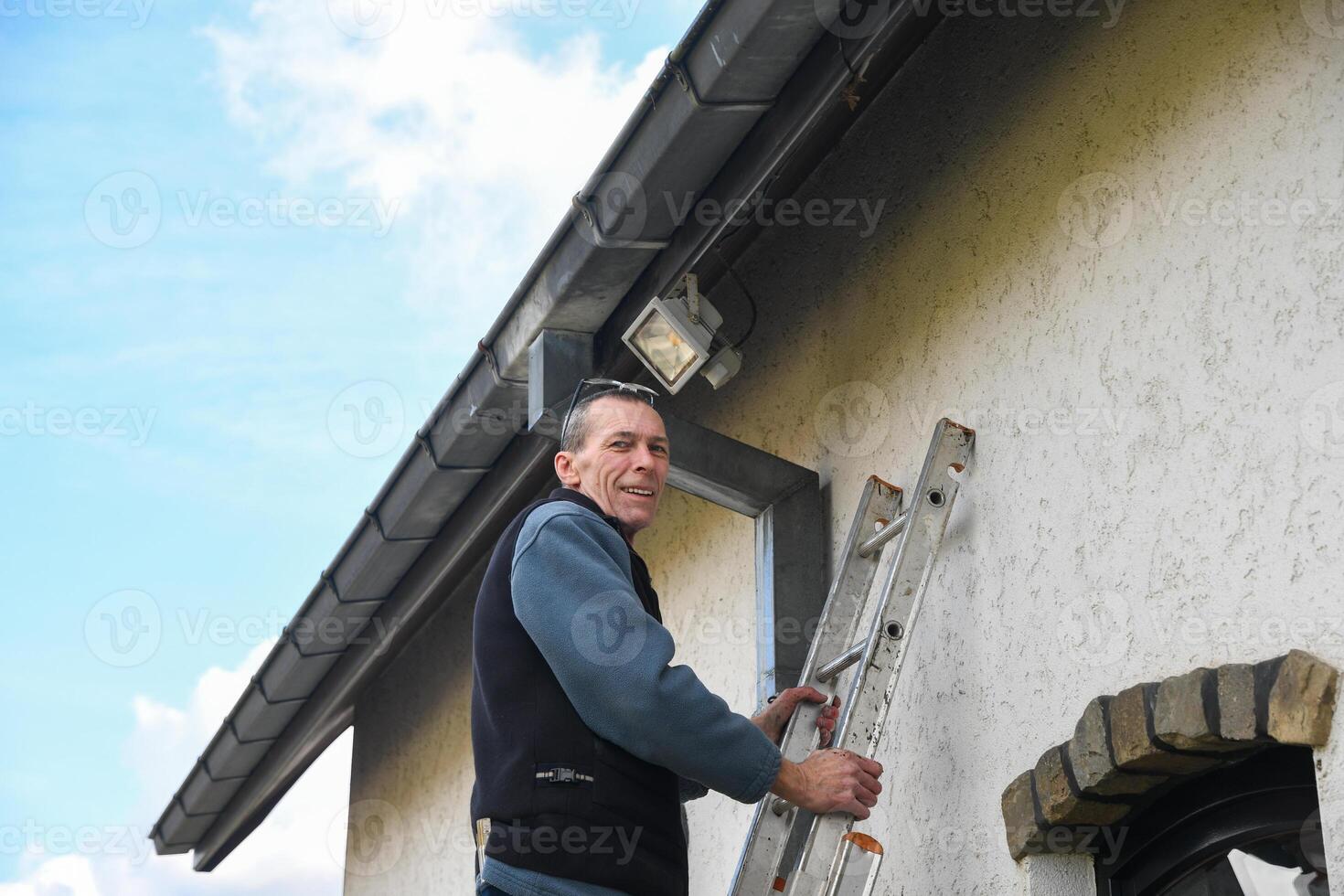 Professional electrician reaches for motion sensor LED floodlight while standing on a ladder, installing it on the facade of a house under a gutter to improve outdoor lighting photo