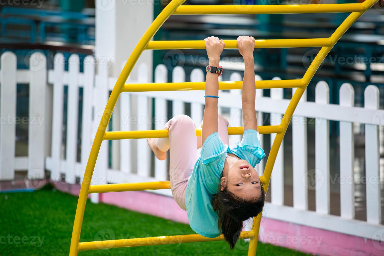 A girl is having fun hanging from an iron ladder in a playground. Little kid smiling and playing climbing joyfully on the playground in the summer park. photo