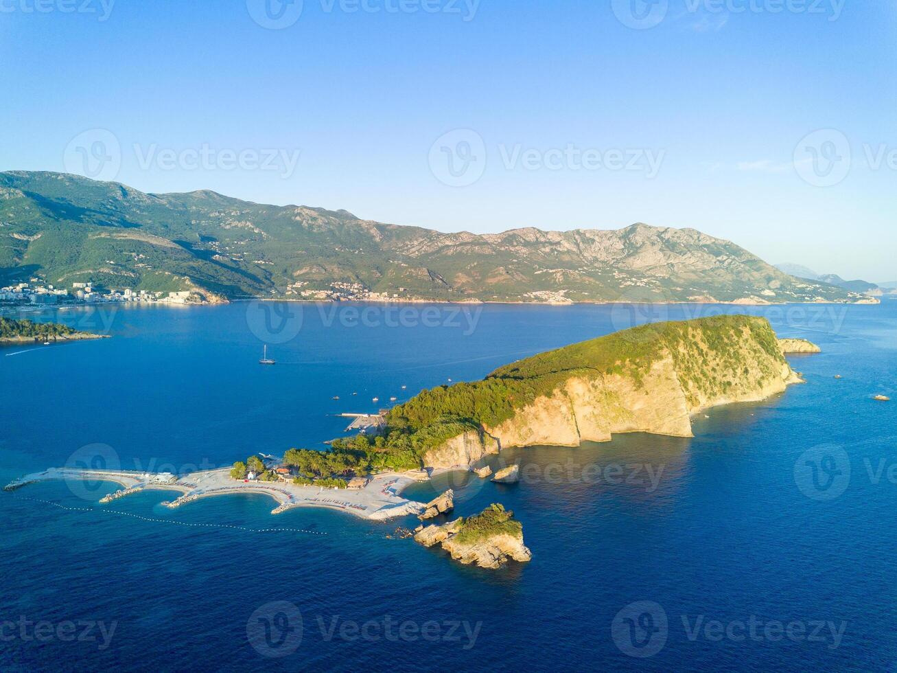 Island of St. Nicholas with vegetation on shores in the Adriatic Sea against the backdrop of coastal towns, the Montenegrin mountains and clear sky photo