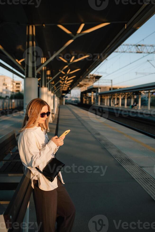 Young woman leaning on bench at railway station platform, using smartphone while waiting for train during sunset, embodying modern urban travel. Woman scroll smartphone, stands on railway platform photo