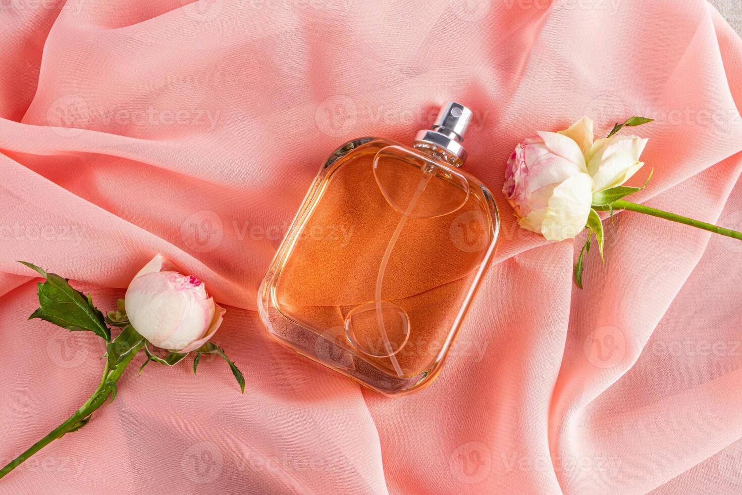 A transparent bottle of cosmetic spray or perfume lies on a chiffon fabric with buds of roses. Top view of a blank mockup of a non-branded bottle. photo