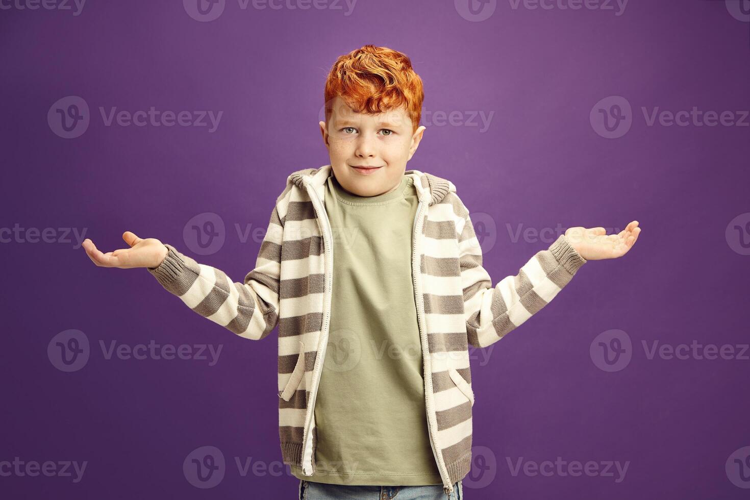 Puzzled and ignorant little red-haired boy shrugs his shoulders, symbolizing lack of knowledge, showing illiteracy wearing in casual clothes standing against violet isolated background photo