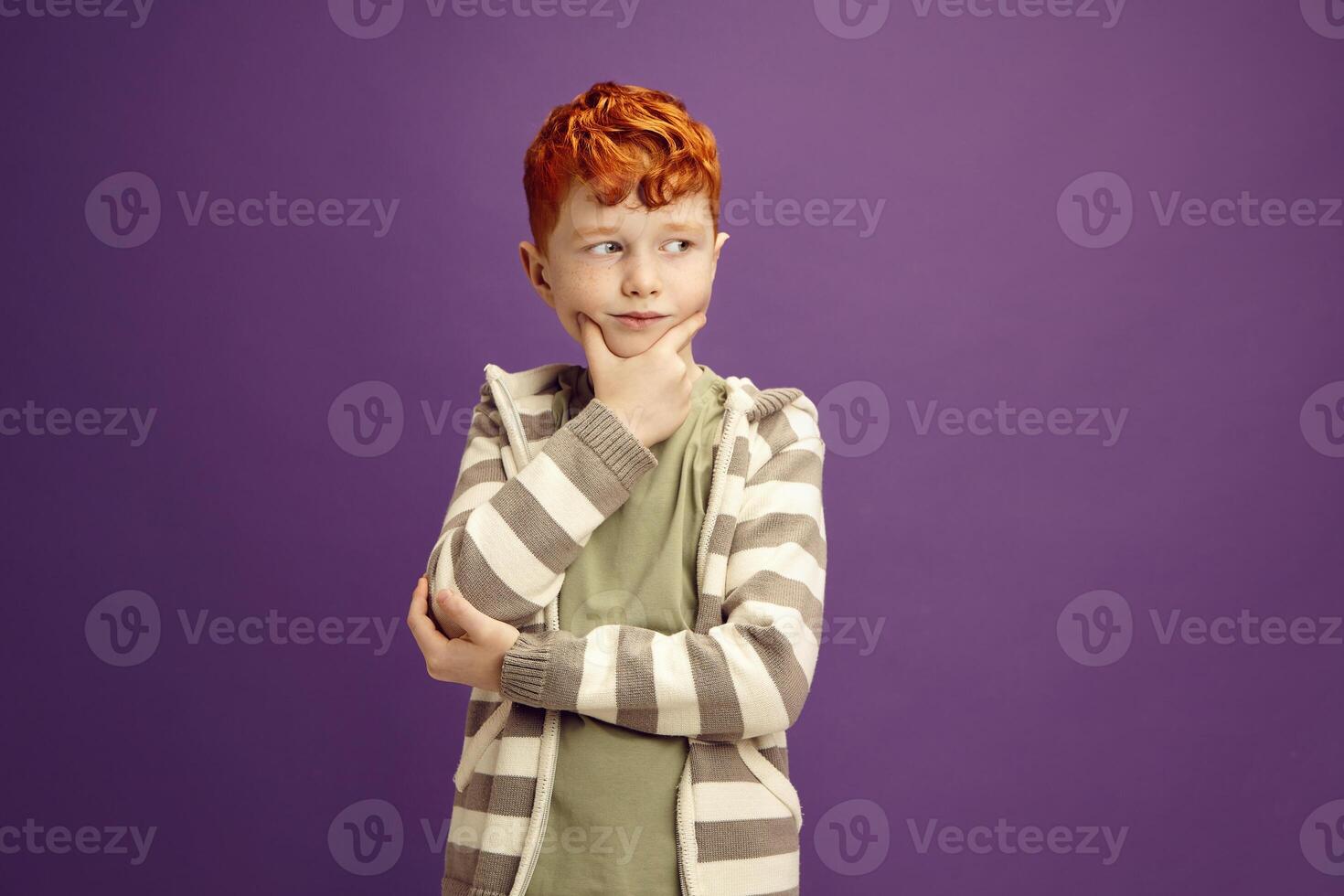 Pensive Red Headed Little Boy Around 7-8 years Puzzled Standing Keeping Hand to Cheek with Thoughtful Face Expression Looking Aside, Wearing in Casual Clothes Standing on Isolated Violet Background photo