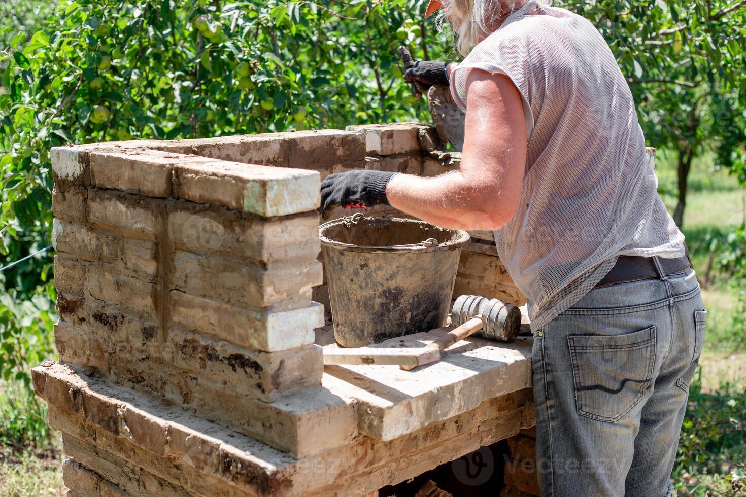 A man builds a brick barbecue structure outdoors, applying mortar and using a rubber mallet, demonstrating the construction process and craftsmanship photo