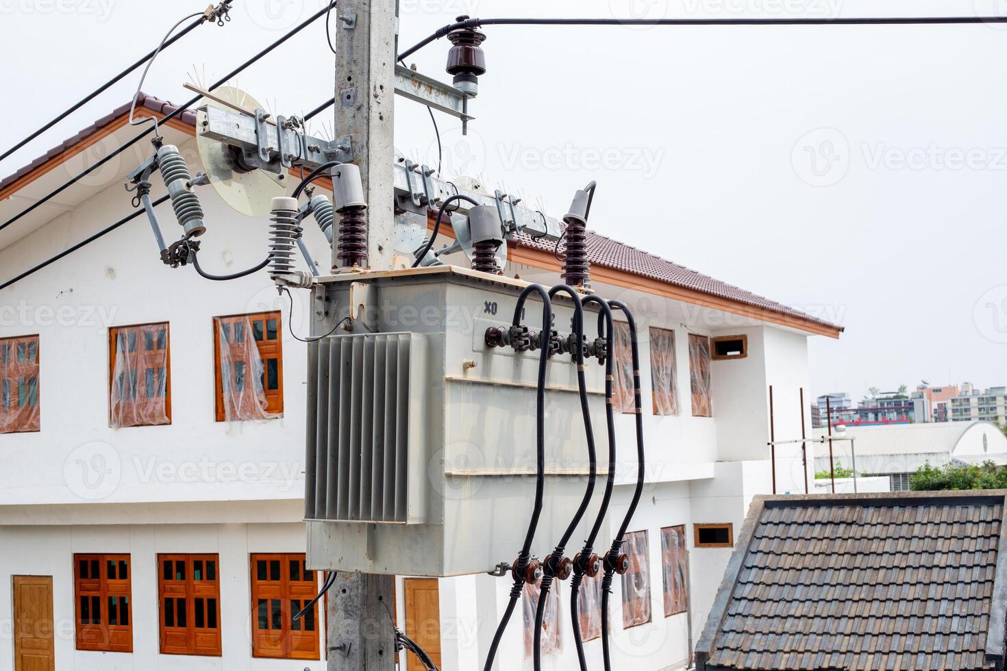 A distribution transformer mounted on a power line pole is located among residential buildings, providing electricity to the entire area photo
