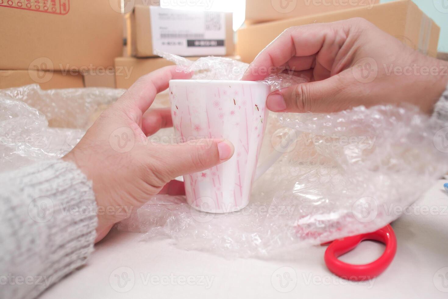 Hands unboxing or packing a ceramic mug with bubble wrap, showing fragile item care, shipping, surrounded by other delivery packages. photo