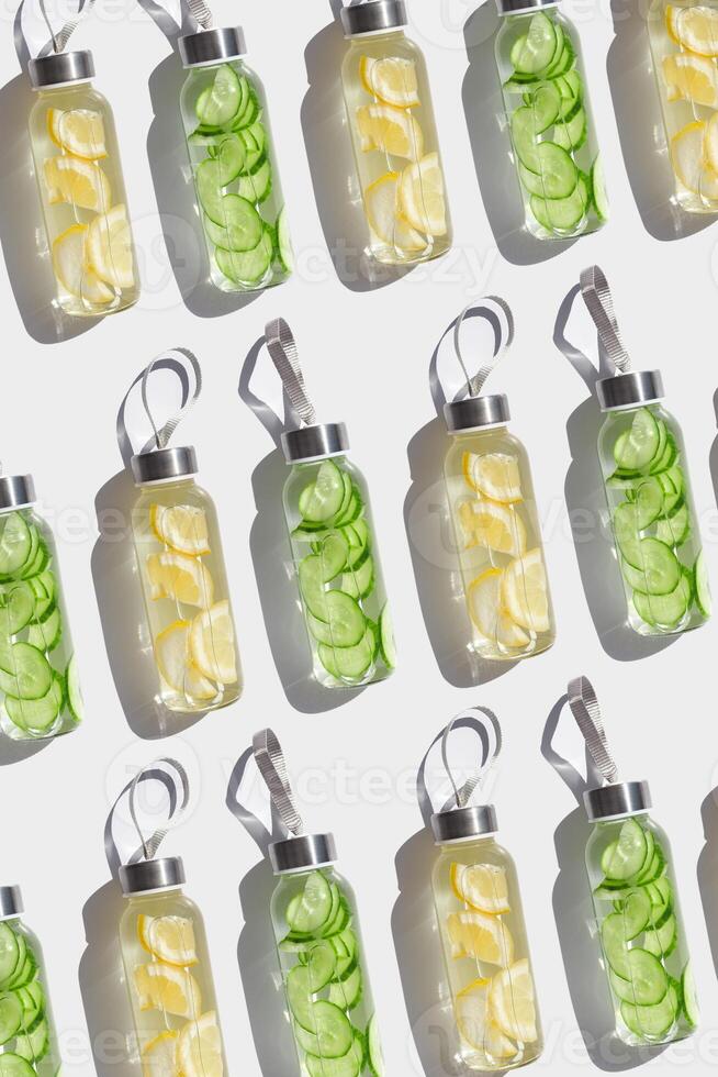 Top view glass bottles, water drink detox with lemon and cucumber at sunlight on white background, geometric pattern. Infused water with slices fruit or vegetable. Aesthetic photo with drinks
