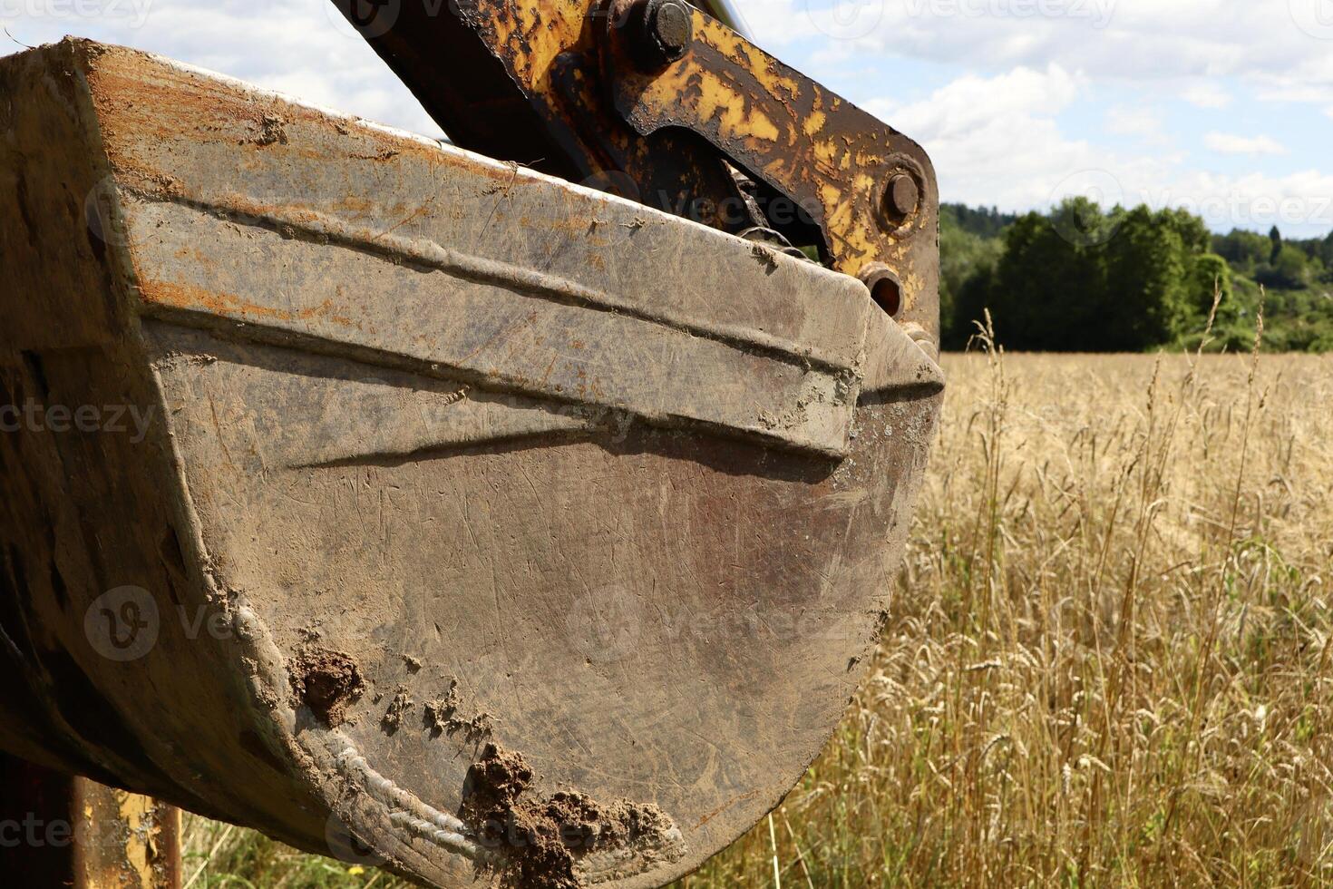 A close-up of a weathered and slightly rusted excavator bucket with traces of soil, set against a backdrop of a ripe rye field and a distant tree line under a partly cloudy sky. photo