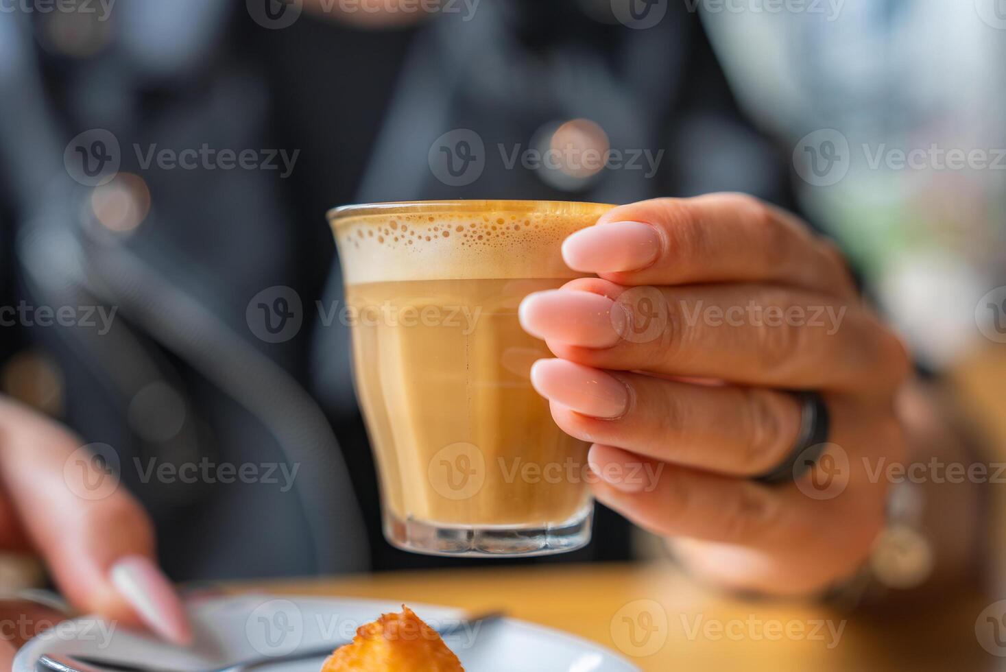 A hand with manicured nails holds a small glass of coffee with foam. A plate with a pastry is visible, set in a softly blurred indoor cafe, possibly in Utrecht. photo