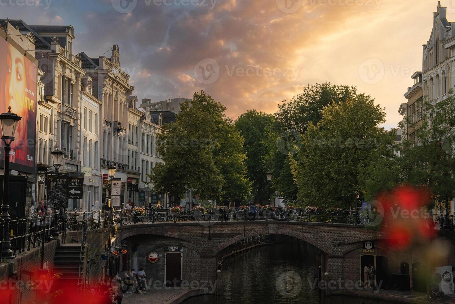 A canal in Utrecht, Netherlands, with a stone bridge, traditional Dutch buildings, trees, and people dining. Red flowers and sunset hues enhance the scene. photo