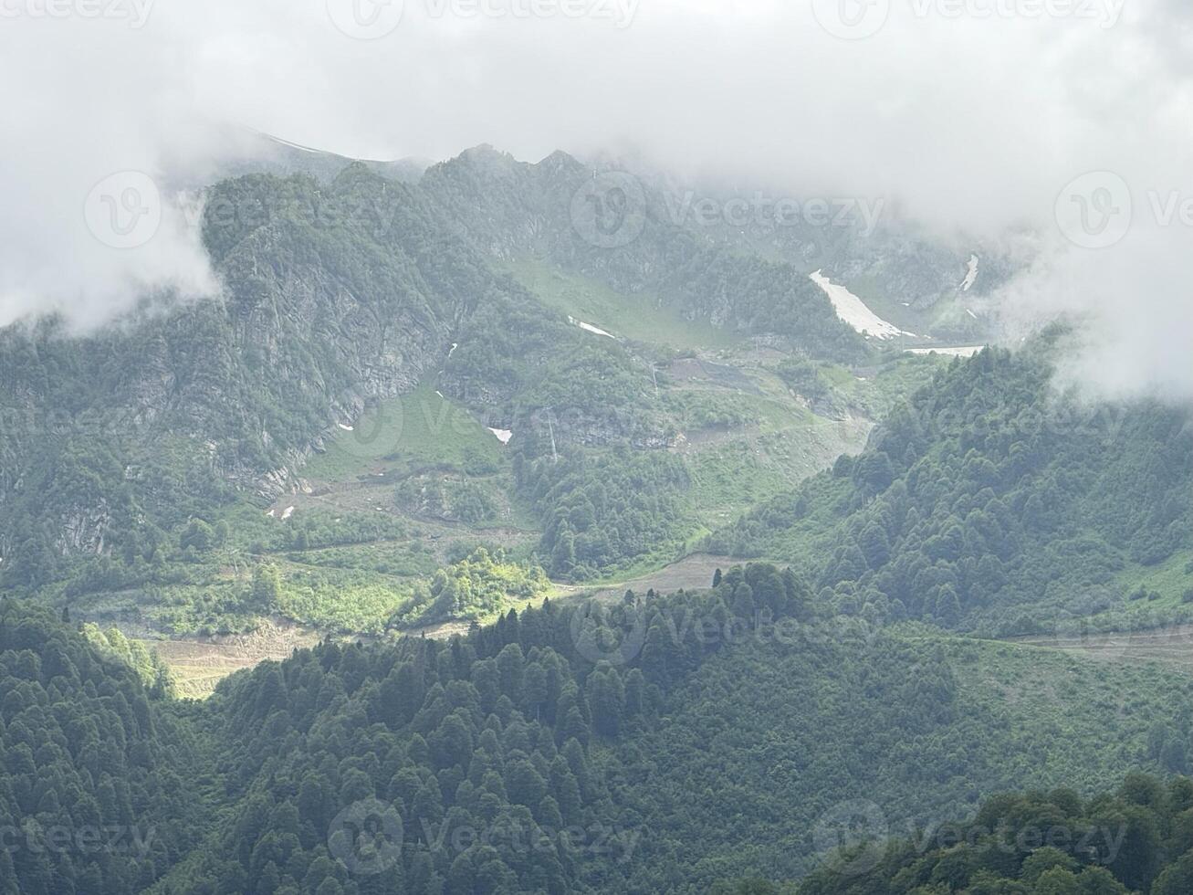 Low clouds are swirling around the peaks of lush green mountains, creating a breathtaking scene of untouched nature, where the remaining snow patches highlight the rugged slopes photo