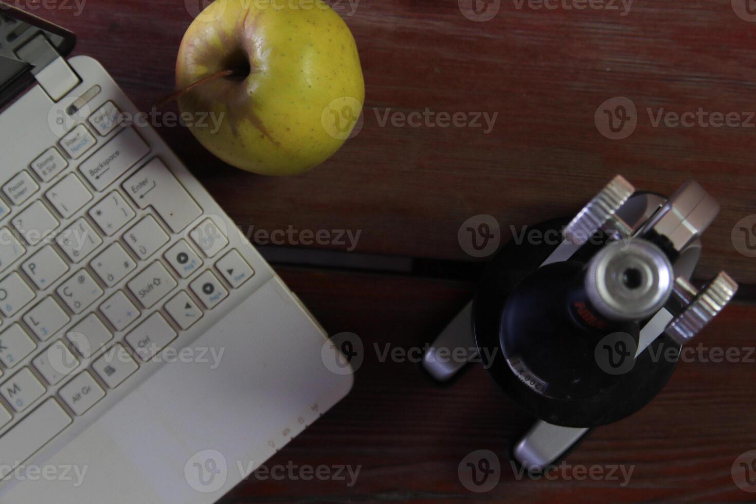 There is a microscope and a laptop on the wooden table. The emphasis is on the apple. photo