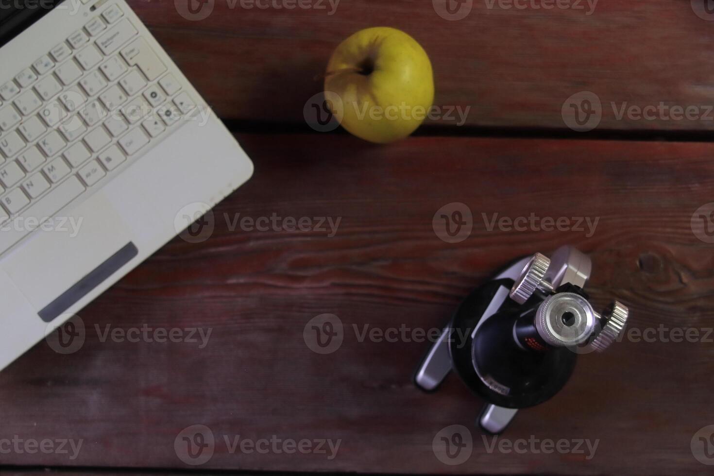 There is a microscope, a laptop and a green apple on the wooden table. photo