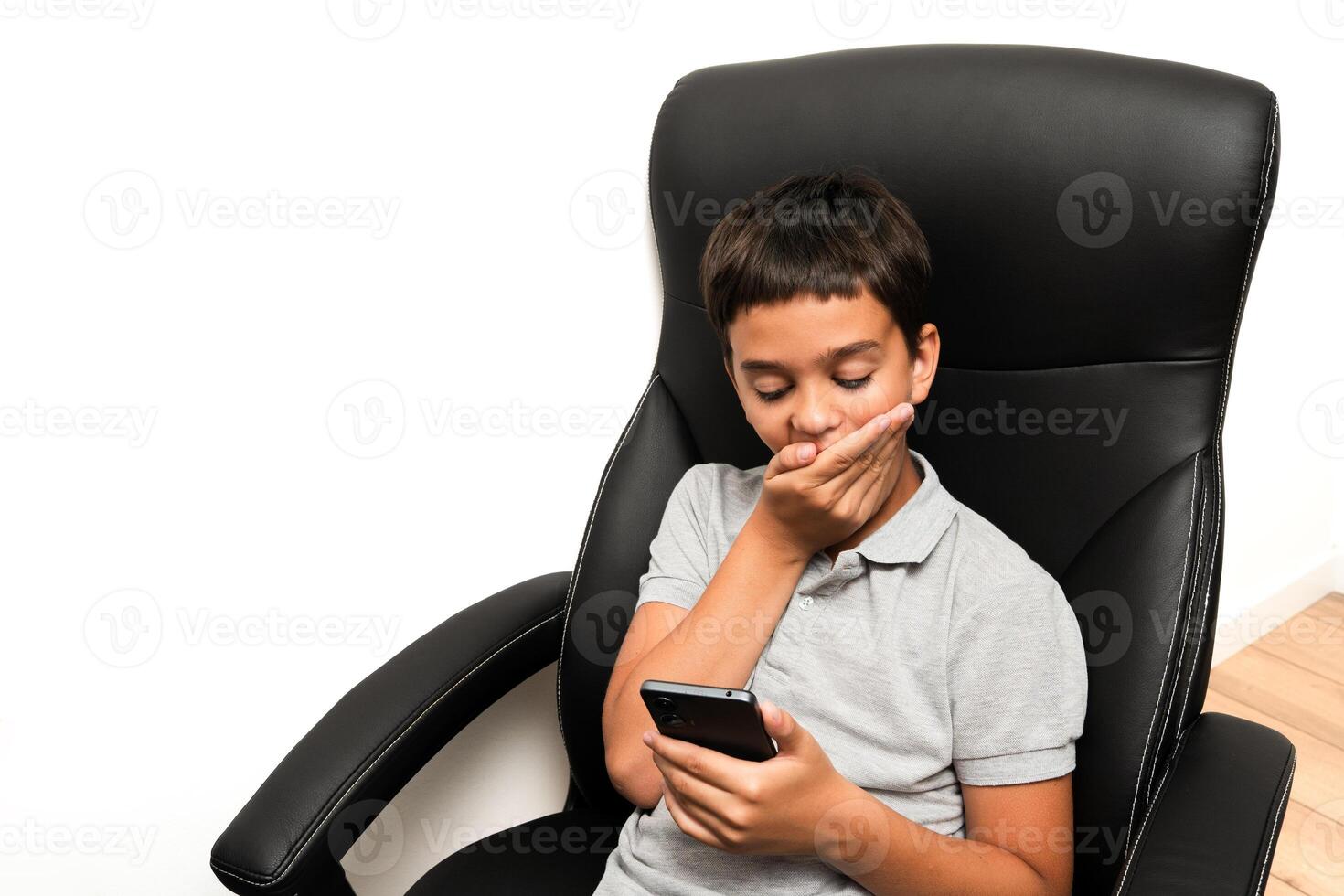 A young boy in a gray polo shirt sits on a black chair, holding a smartphone and covering his mouth in surprise, isolated on a white background. Suitable for concepts related to technology use, digital communication, and emotional reactions in children. photo