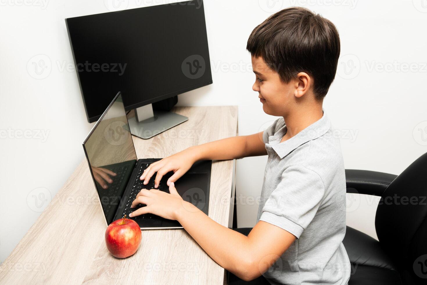 Young boy in a gray polo shirt sits at a wooden desk using a laptop, with an apple and a monitor nearby. Suitable for illustrating remote learning, home study, or technology use by children. photo