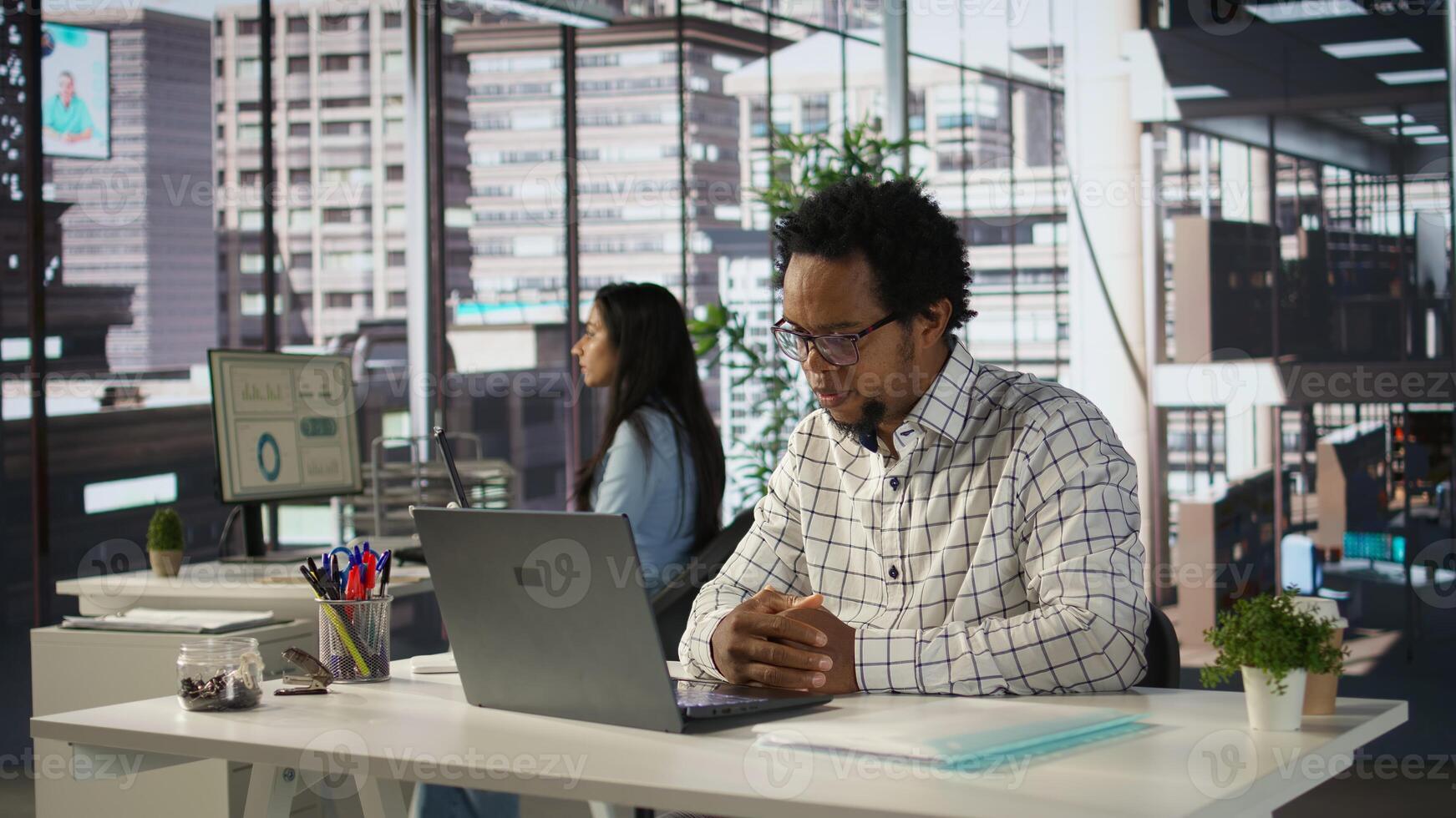 Black man employee joins a telecommuting session at his office desk, multitasking between project management and business strategies. Productivity and collaboration across departments. Camera B. photo