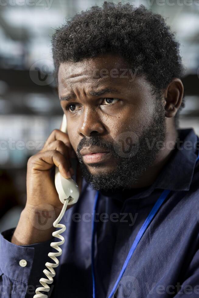 Black corporate employee engages in a landline phone call at the office, managing a project briefing and discussing tasks completion with the supervisor. Multitasking for productivity. photo