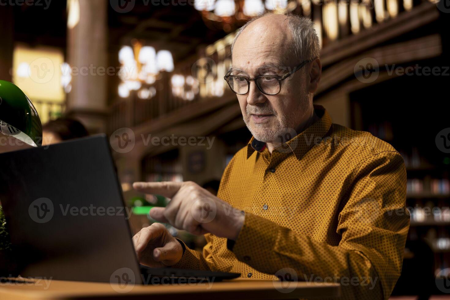 Elderly wise college student interacting with professor at webinar on laptop, studying in a library and combining modern telecommunication with classic academic research. Seminar lesson. photo