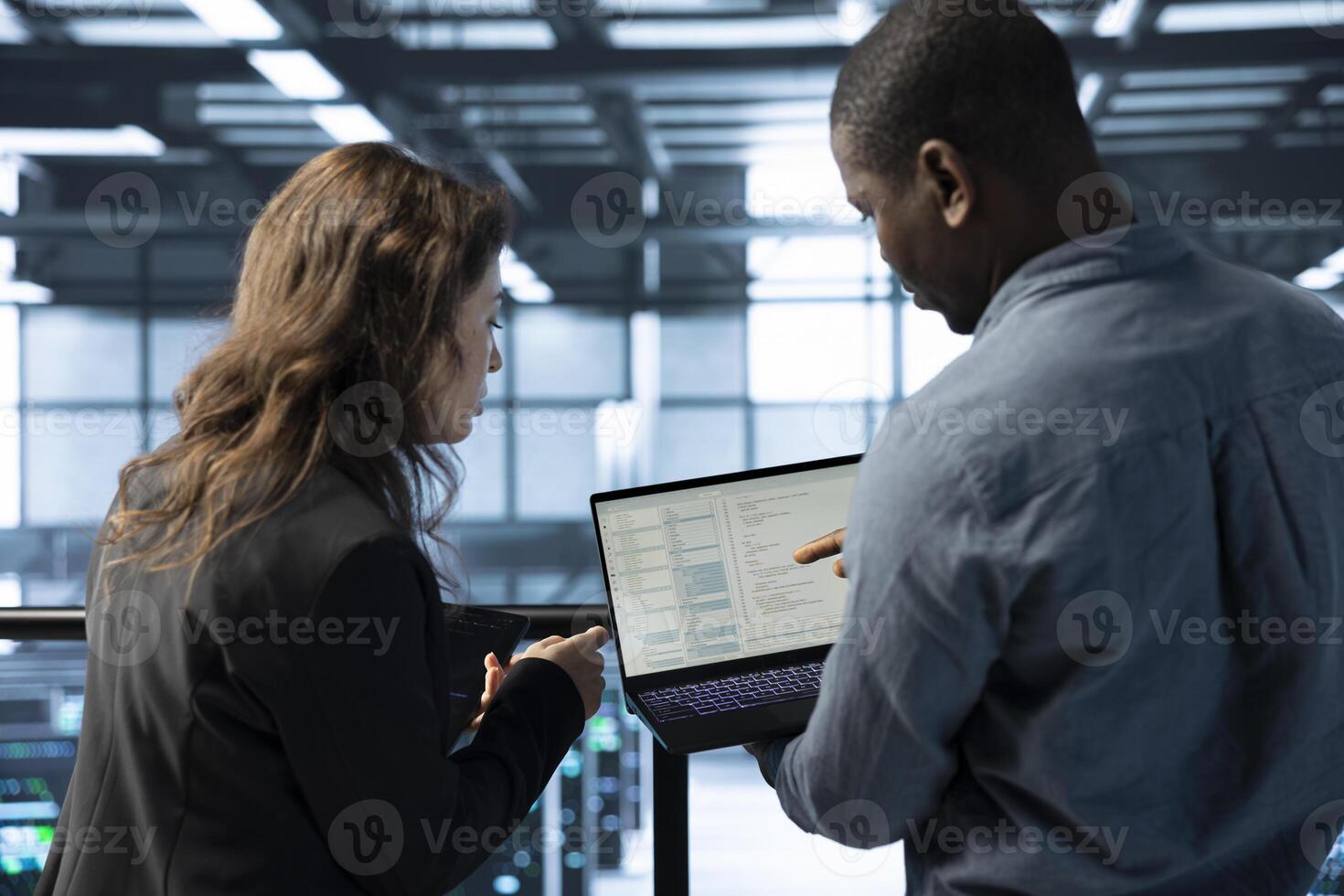 Programmers in data center workspace coding on laptop, analyzing metrics, optimizing storage volumes. Teamworking IT professionals in server hub using notebook, chatting, ensuring seamless operations photo