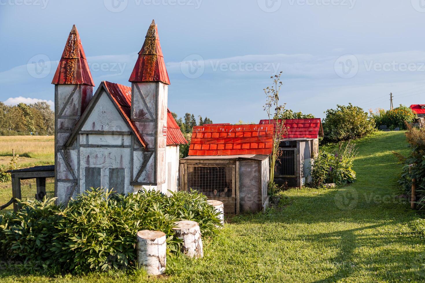 Outdoor scene with a whimsical castle like structure with red pointed roofs, rabbit cages nearby, green grass, plants, tree stumps, and open fields. photo