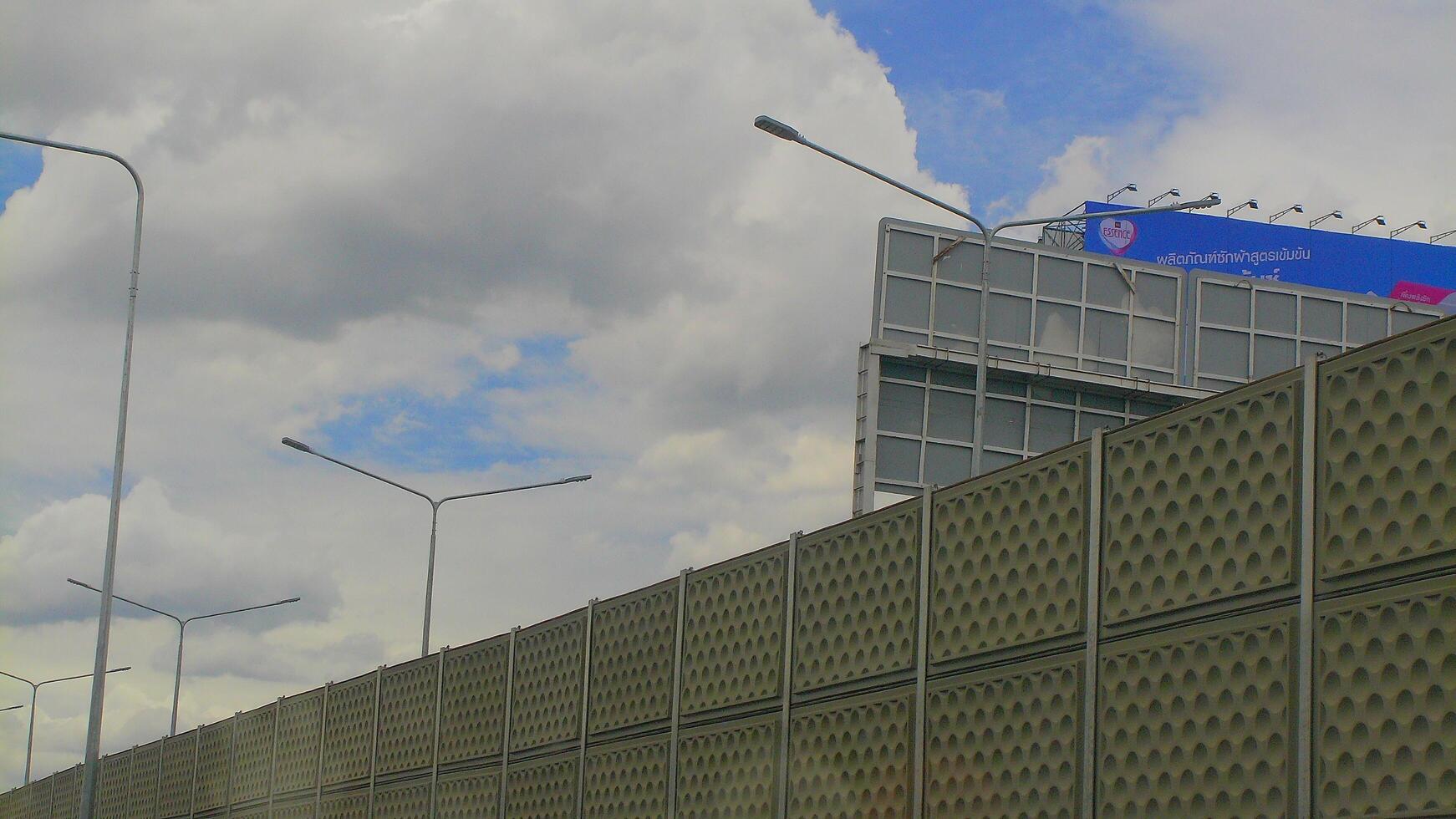 A highway sound barrier and a large billboard in Bangkok, seen from a moving vehicle under partly cloudy skies, illustrating urban noise control and advertising. photo