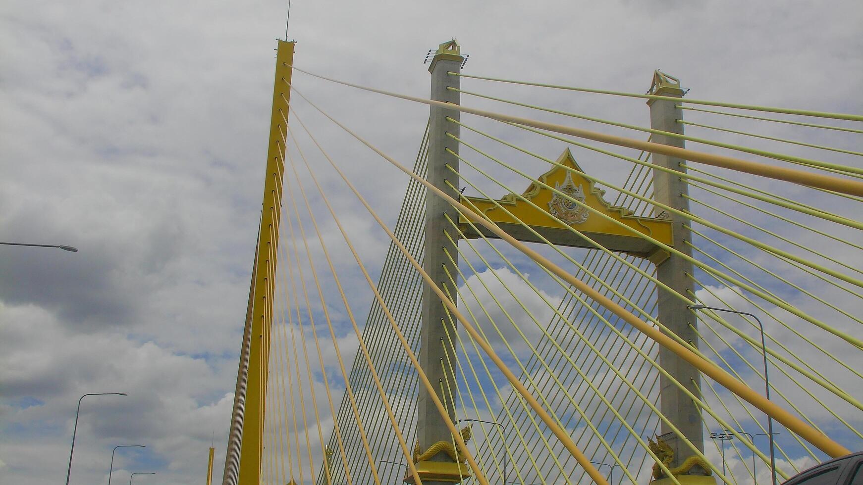 Towering structure of Bhumibol Bridge captured from below, emphasizing the tall suspension pillars and radiating yellow cables beneath an overcast sky. photo
