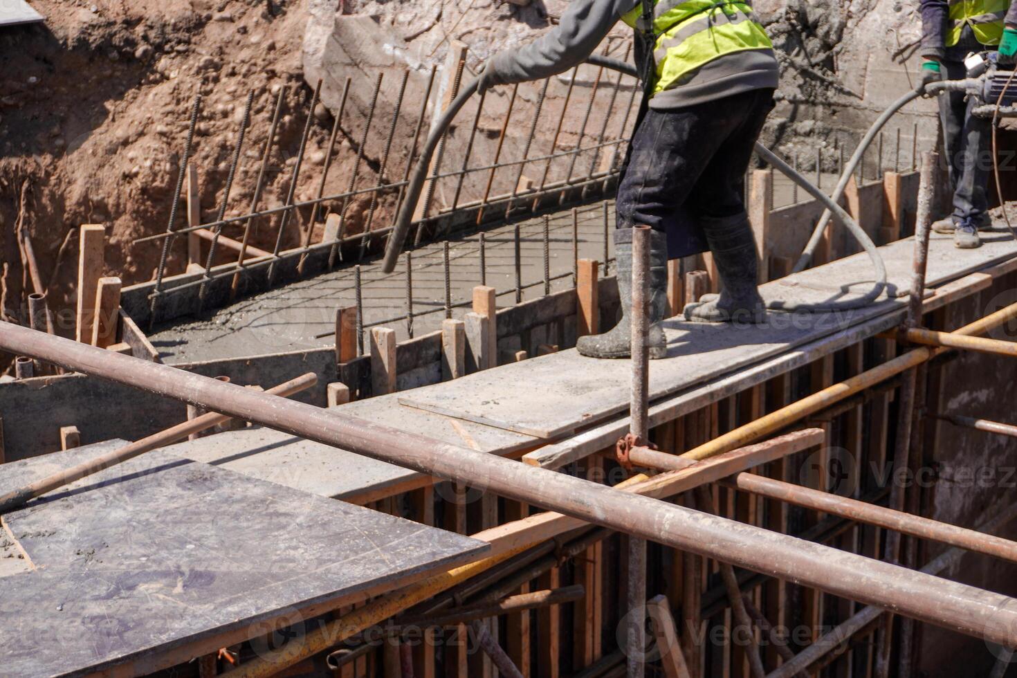 Workers in safety vests pour concrete into wooden forms, with metal rebar protruding. The scene shows a partially completed structure with tools nearby. photo