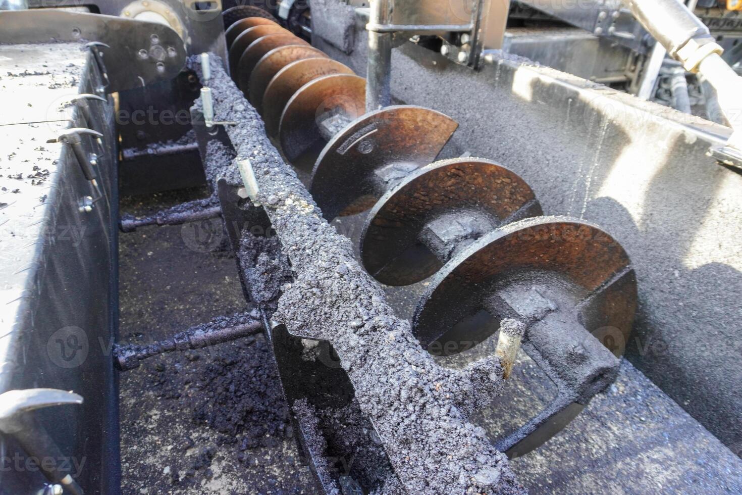 Close-up of an asphalt mixing machine's spiral blades, showcasing rusted metal surfaces, dark asphalt residue, and mechanical components in a construction setting. photo