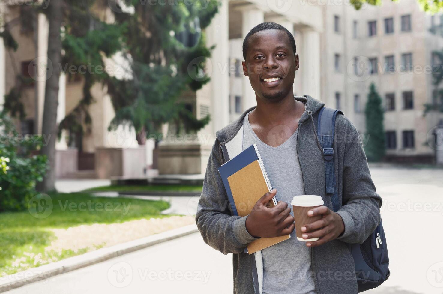 Smiling african-american student with books and take away coffee outdoors, having rest in campus. Education concept, copy space photo