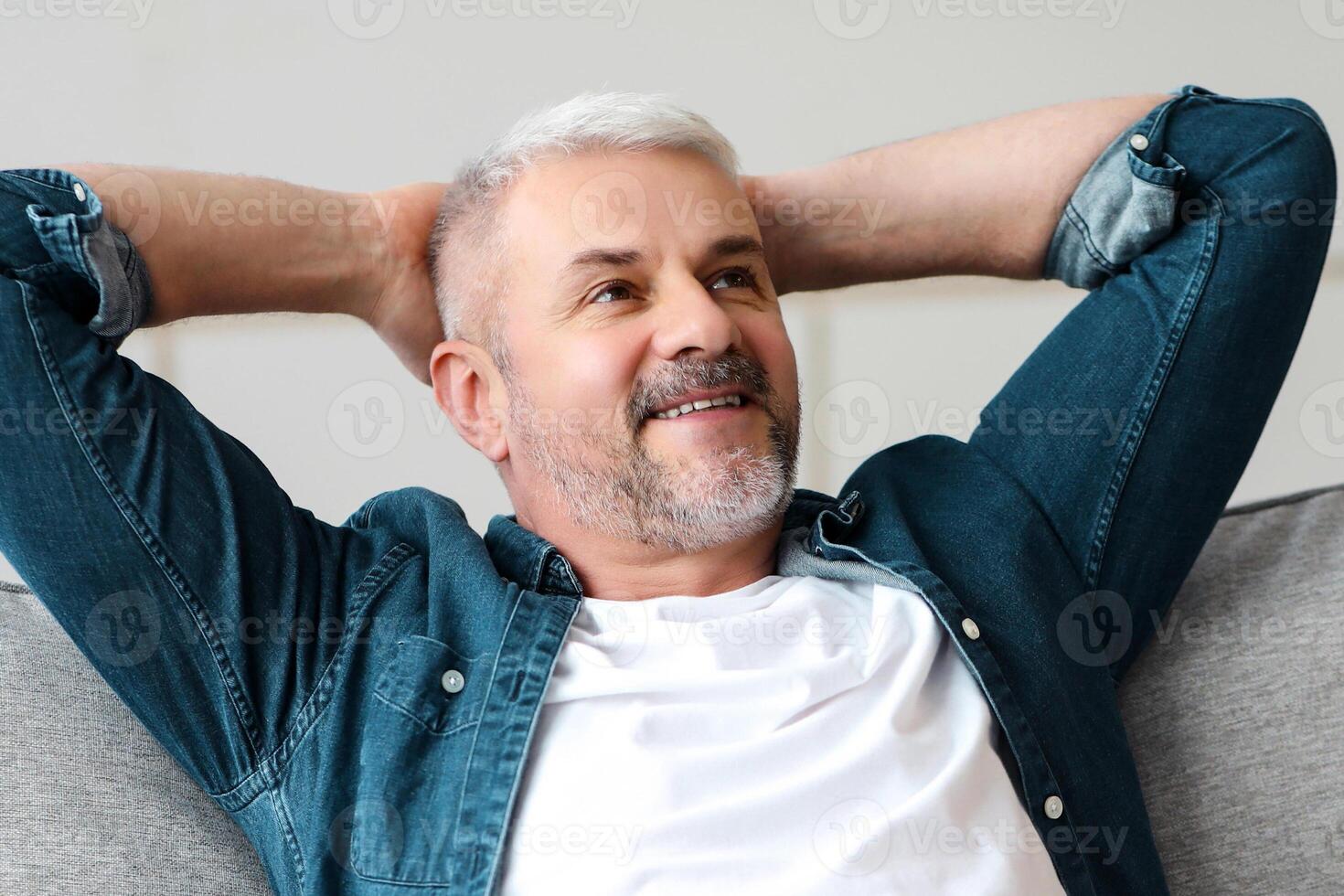 Happy bearded senior man in casual clothes relaxing on couch at home, looking at copy space. Relaxed matured man having break on sofa in living room, dreaming about prosperous life photo