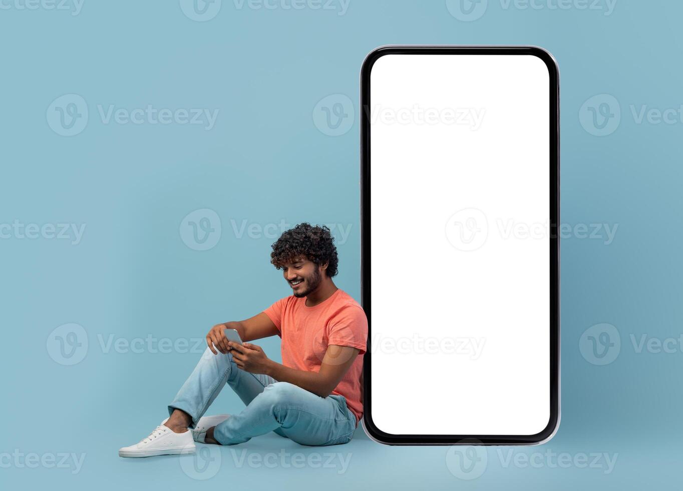 Mobile app concept. Relaxed curly young indian man using cell phone on blue studio background, sitting by big mobile phone with white empty screen, full length shot, panorama with copy space, mockup photo