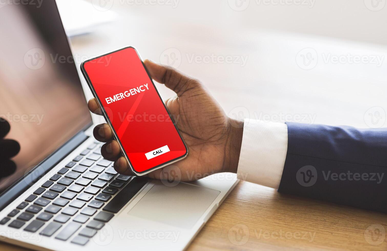 Safety Concept. Male hand holding smart phone with emergency number on red screen over office background and laptop photo