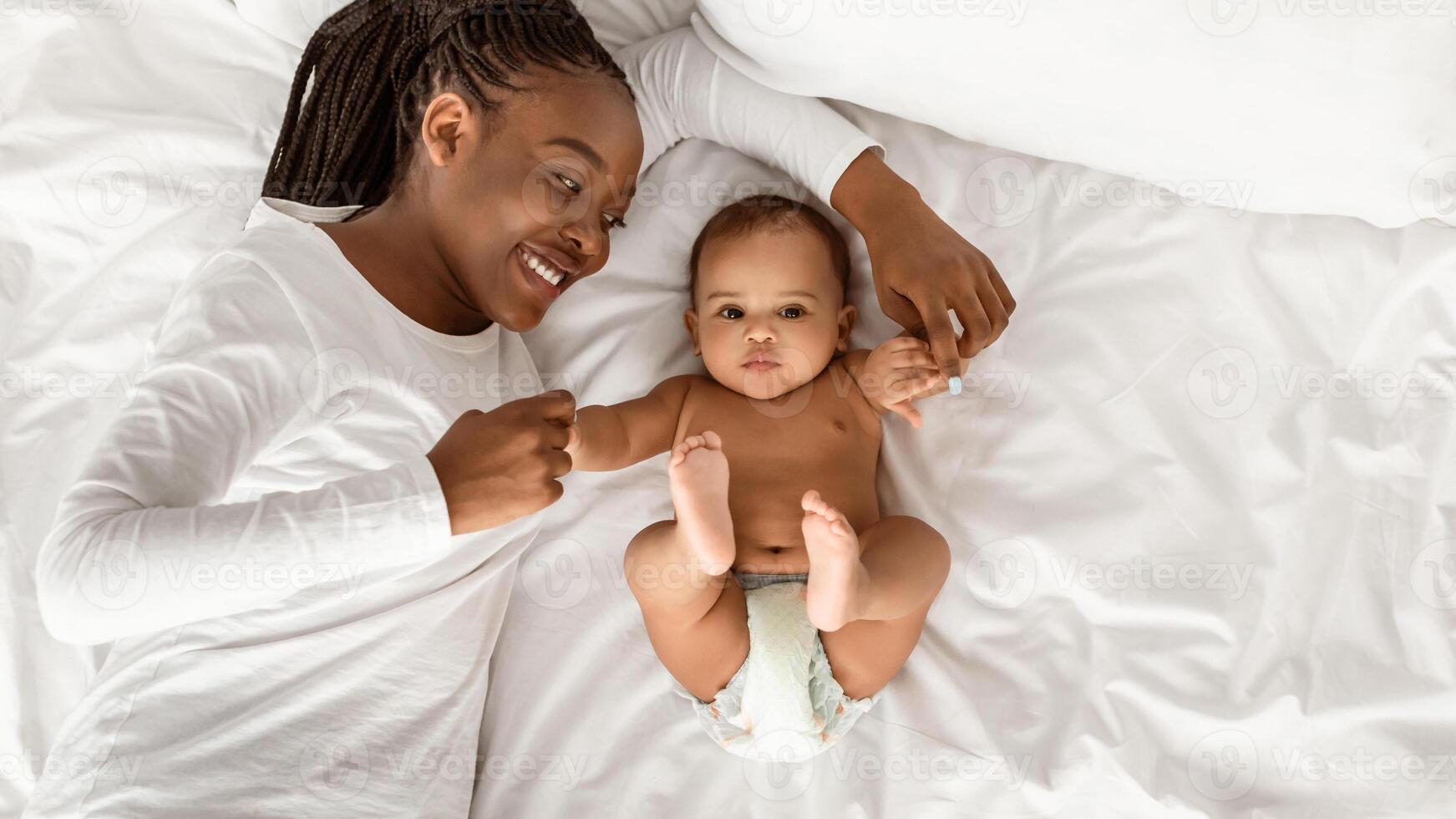 Happy Maternity. Above top high angle view portrait of positive African American woman lying in bed and playing with her cute little baby, touching infant's arms, spending time on white bedsheets photo