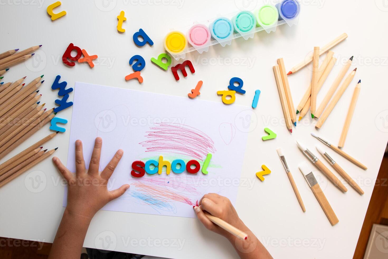 Child drawing with colorful letters, crayons, and paints on white table photo