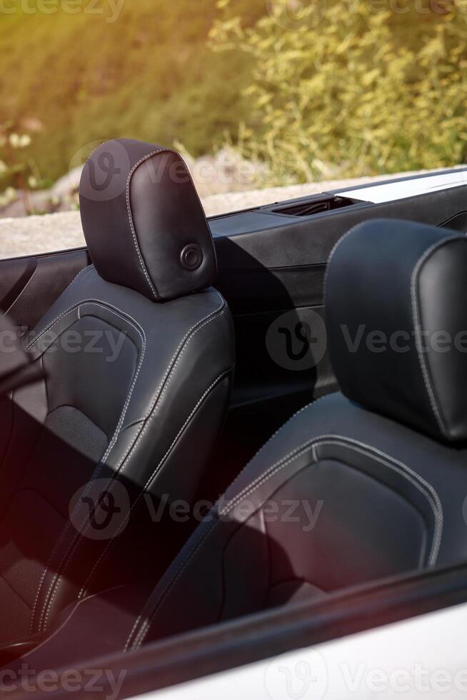 Convertible interior in bright daylight showing black leather seats with crisp white stitching, detailed headrests, vibrant greenery backdrop. photo