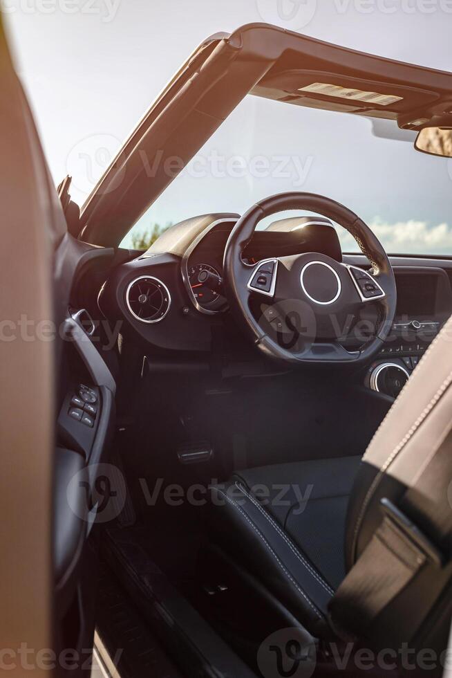 Convertible interior showcasing black leather seats, modern steering wheel, and detailed dashboard with gauges and buttons. Sunlit daytime scene, no people. Ideal for automotive and travel ads. photo