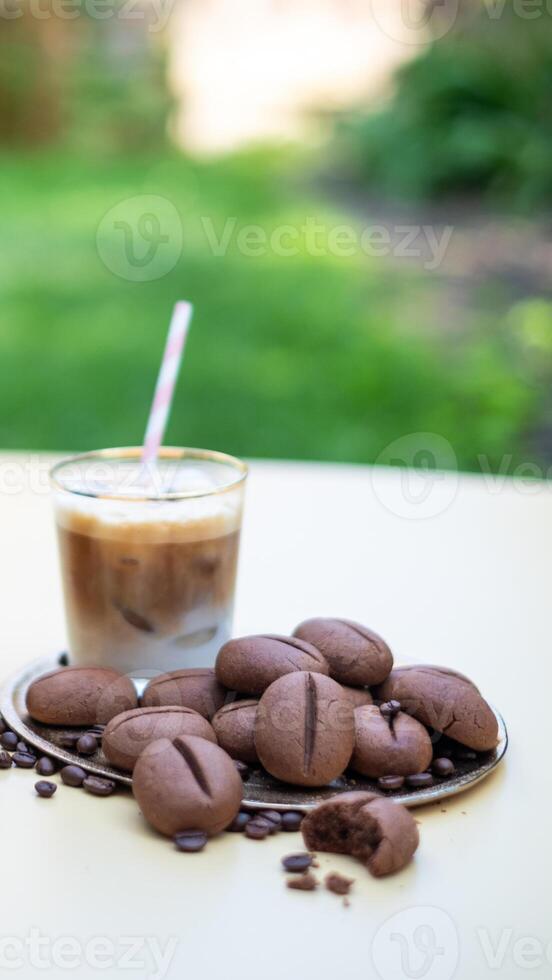 Iced coffee in a glass with a straw beside a plate of chocolate cookies and scattered coffee beans on a light table, creating a delightful dessert scene photo