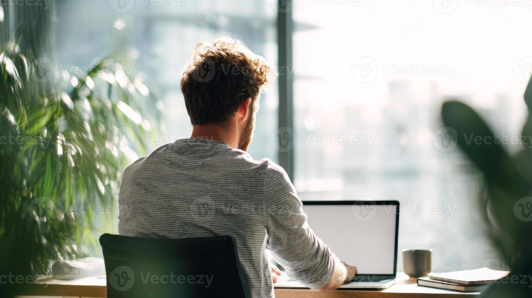 Young man working on laptop in modern office with bright sunlight filtering through window and indoor plants in background photo