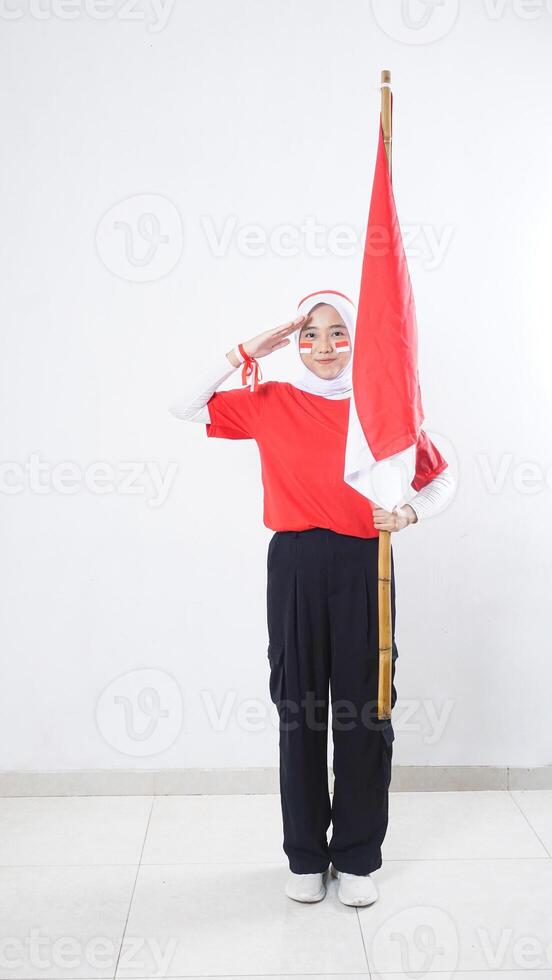 Muslim Asian girl celebrate Indonesia independence day in red and white outfit by holding Indonesian flag and showing salute with hand raised to eyebrow isolated on white background photo