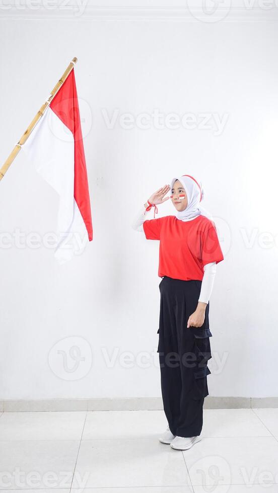 Muslim Asian girl celebrate Indonesia independence day in red and white outfit by showing salute to Indonesian flag isolated on white background photo