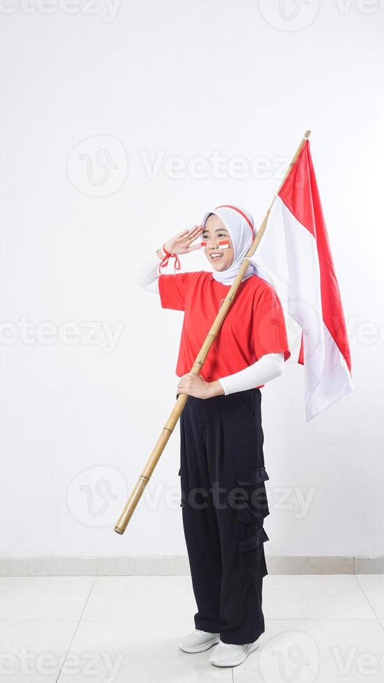 Happy muslim Asian girl celebrate Indonesia independence day in red and white outfit by holding Indonesian flag and doing salute with right hand raised to eyebrow isolated on white background photo