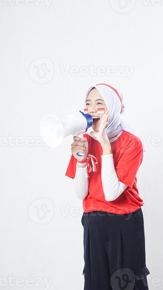 Excited muslim Asian girl celebrate Indonesia independence day in red and white outfit by holding and speaking through megaphone isolated on white background photo